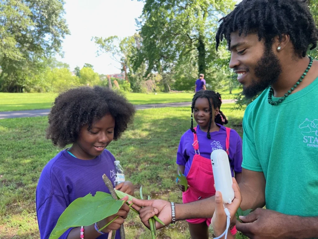 A man in a teal shirt hands a white device to two girls in purple shirts as they examine plant leaves in a sunny park.